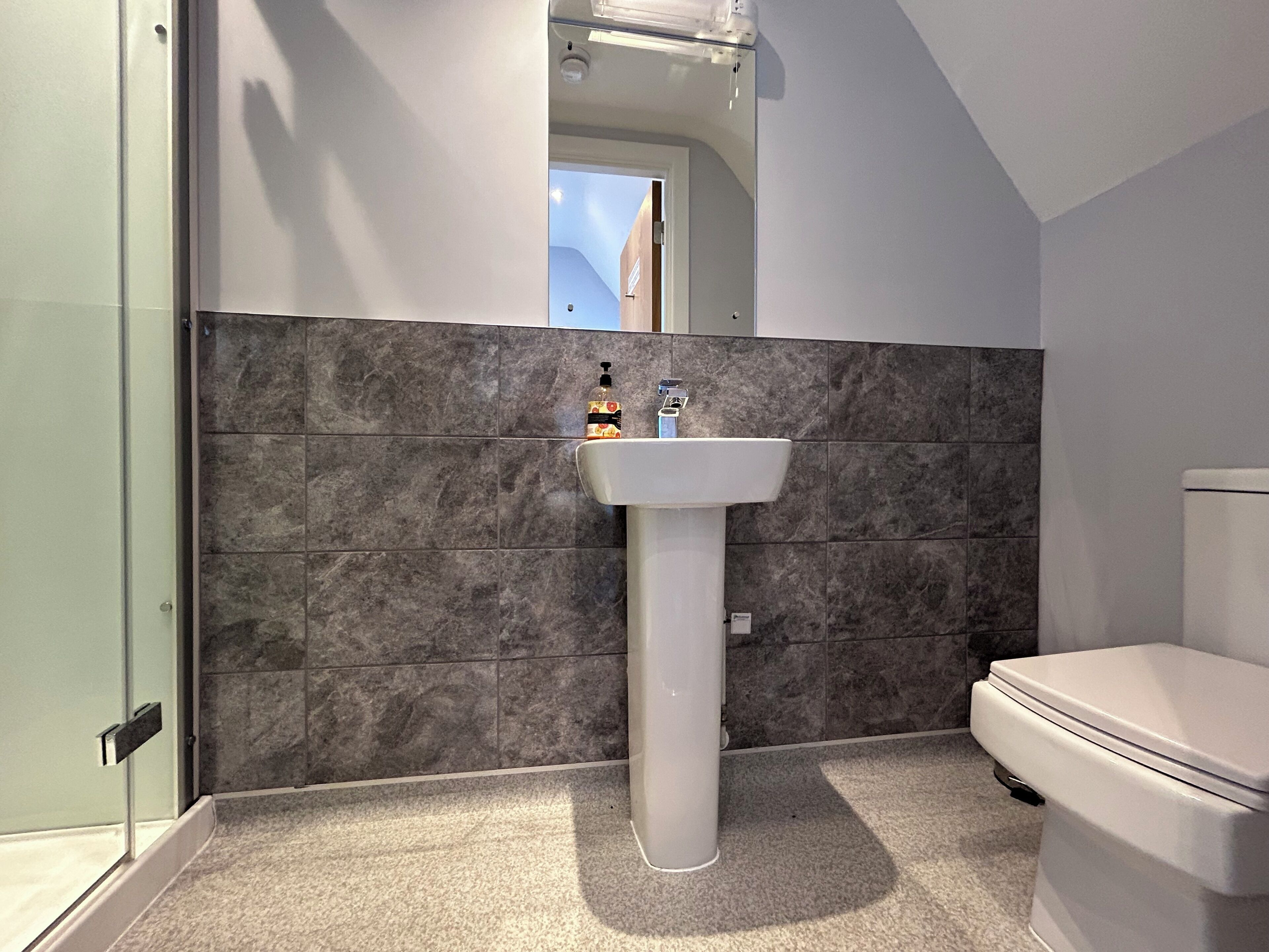Un baño con lavamanos pedestal blanco, baldosas de mármol oscuro, inodoro y cabina de ducha con paredes de vidrio.