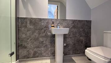 Un baño con lavamanos pedestal blanco, baldosas de mármol oscuro, inodoro y cabina de ducha con paredes de vidrio.