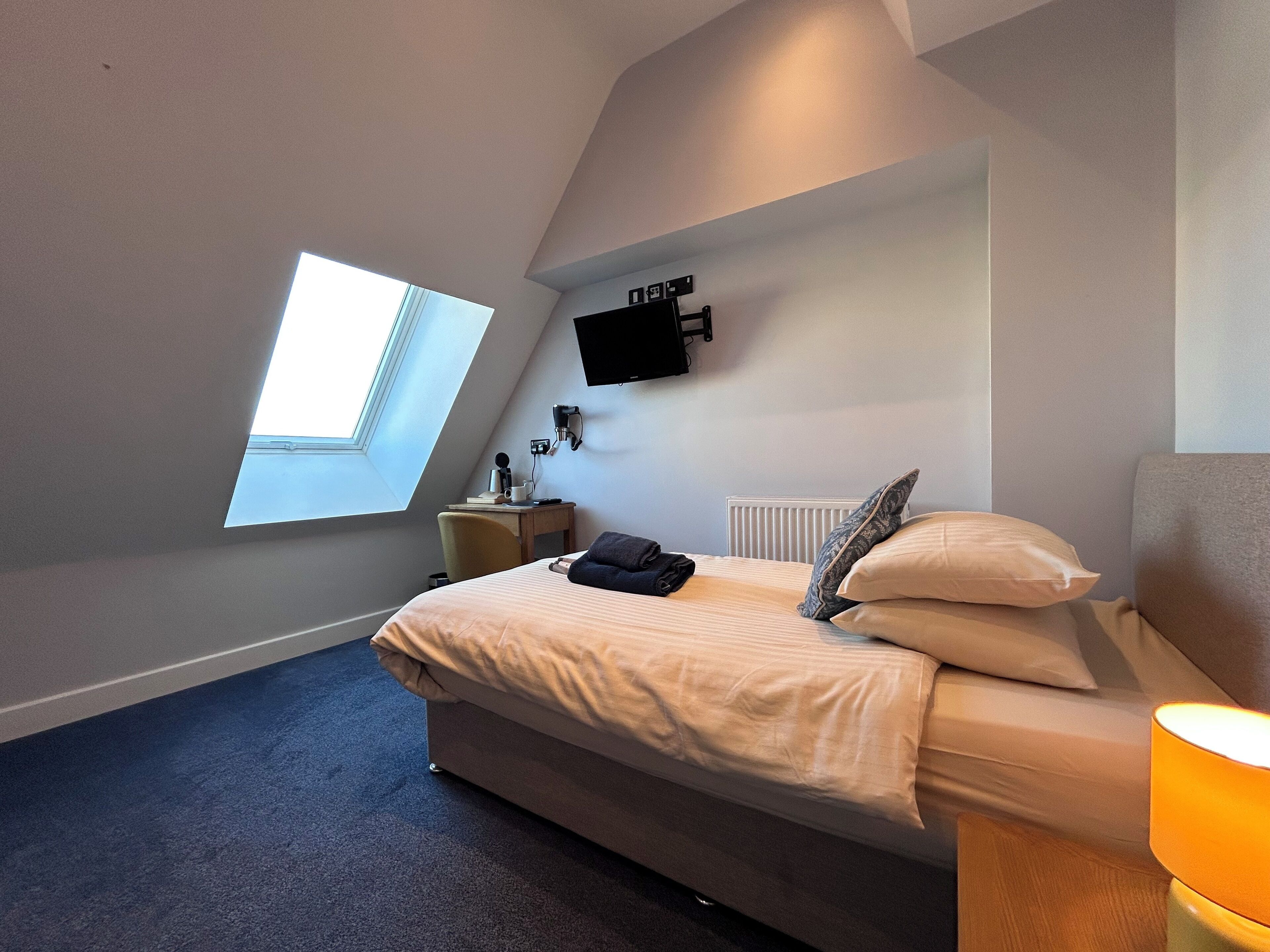 Ein Schlafzimmer mit einem Bett, einem Nachttisch, einer Lampe, einem an der Wand montierten Fernseher und einem Oberlicht.
