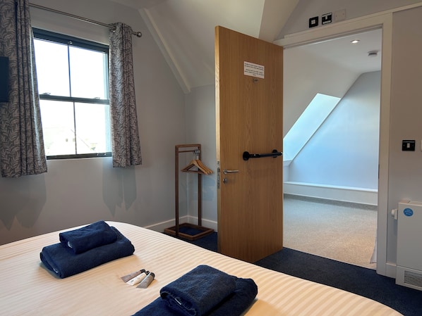 A hotel room with a bed, towels, a wooden door, a coat rack, and a view of a staircase. - Harbour Stays - Ocean Square (Saundersfoot)
