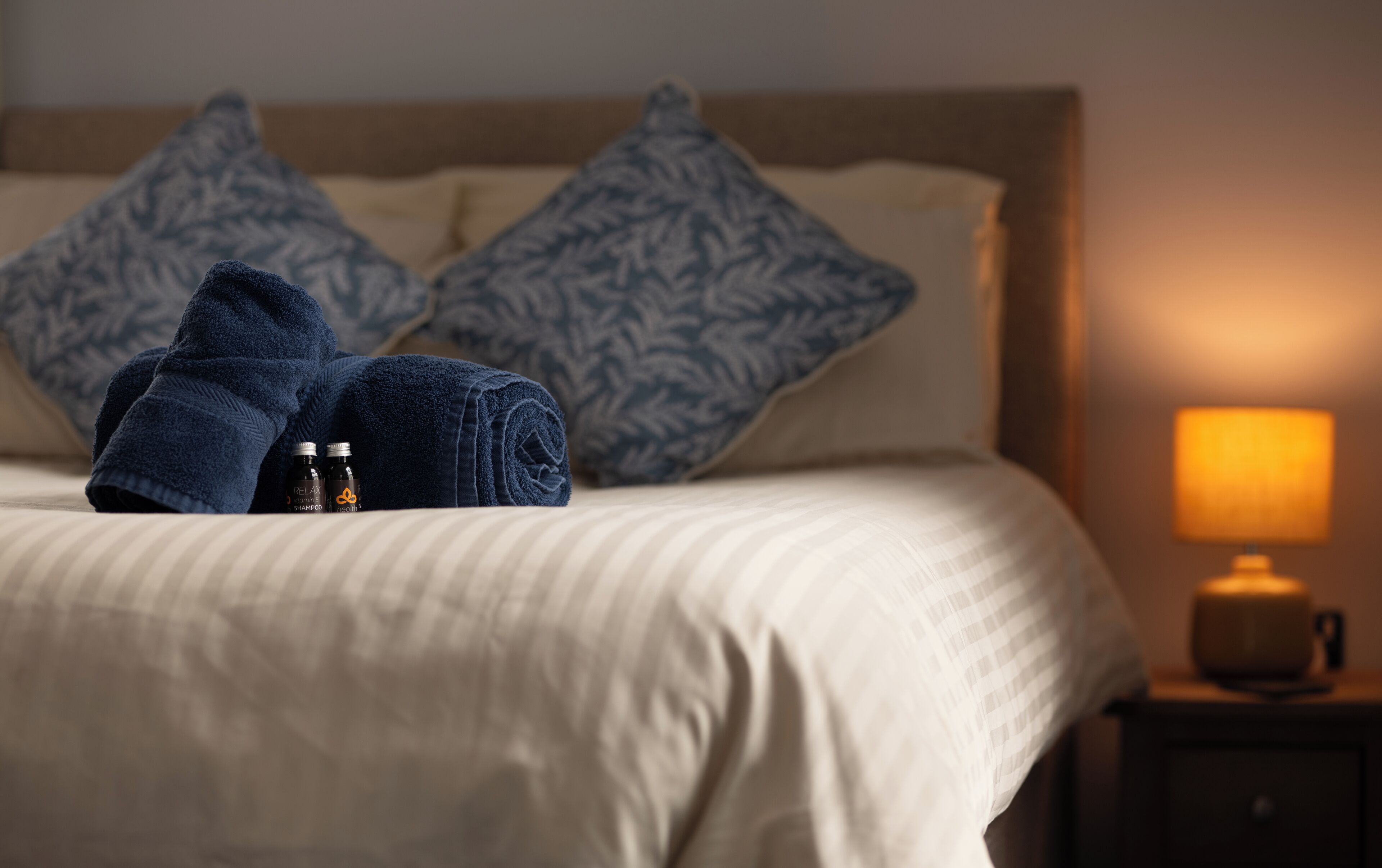 A neatly made bed with a beige bedspread, two folded towels, and two patterned pillows. There are two bottles of shampoo and conditioner on the bed. A bedside lamp is on the nightstand to the right.