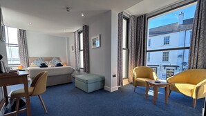 Une chambre d’hôtel comprenant un lit, un bureau, une chaise, un petit pouf et de grandes fenêtres donnant sur la ville.