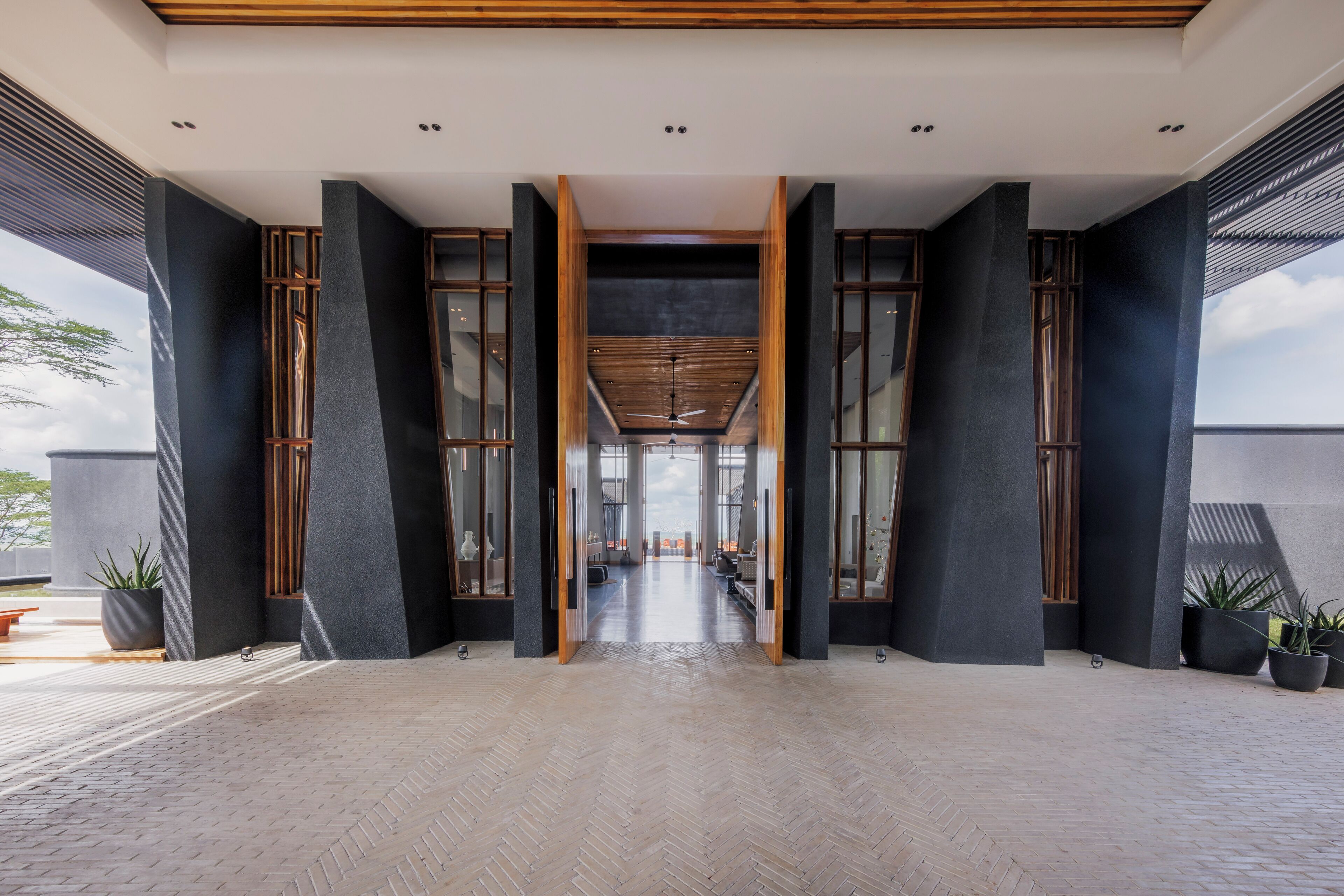 A spacious corridor with a herringbone floor pattern, flanked by dark pillars and wooden beams, leading to a bright interior space.