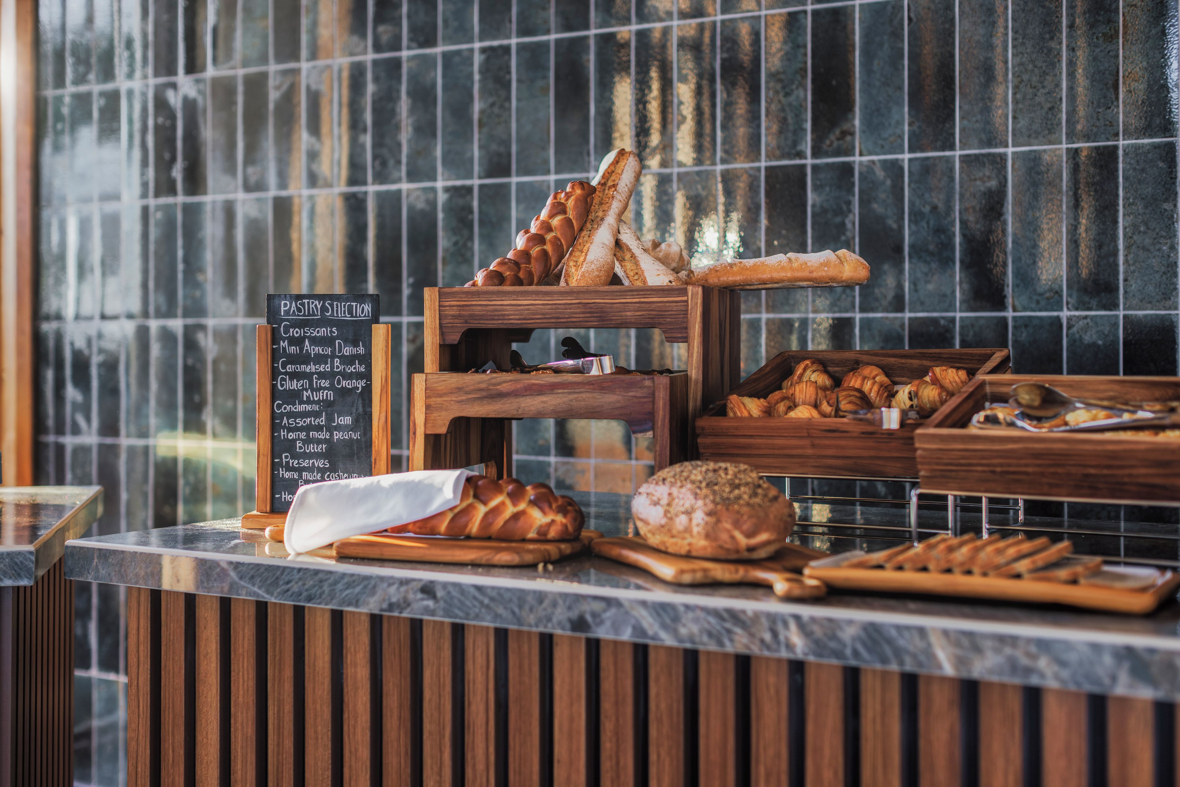 Un expositor de panadería con diferentes panes y pasteles, un menú en una pizarra que enumera los productos, y una pared de azulejos al fondo.