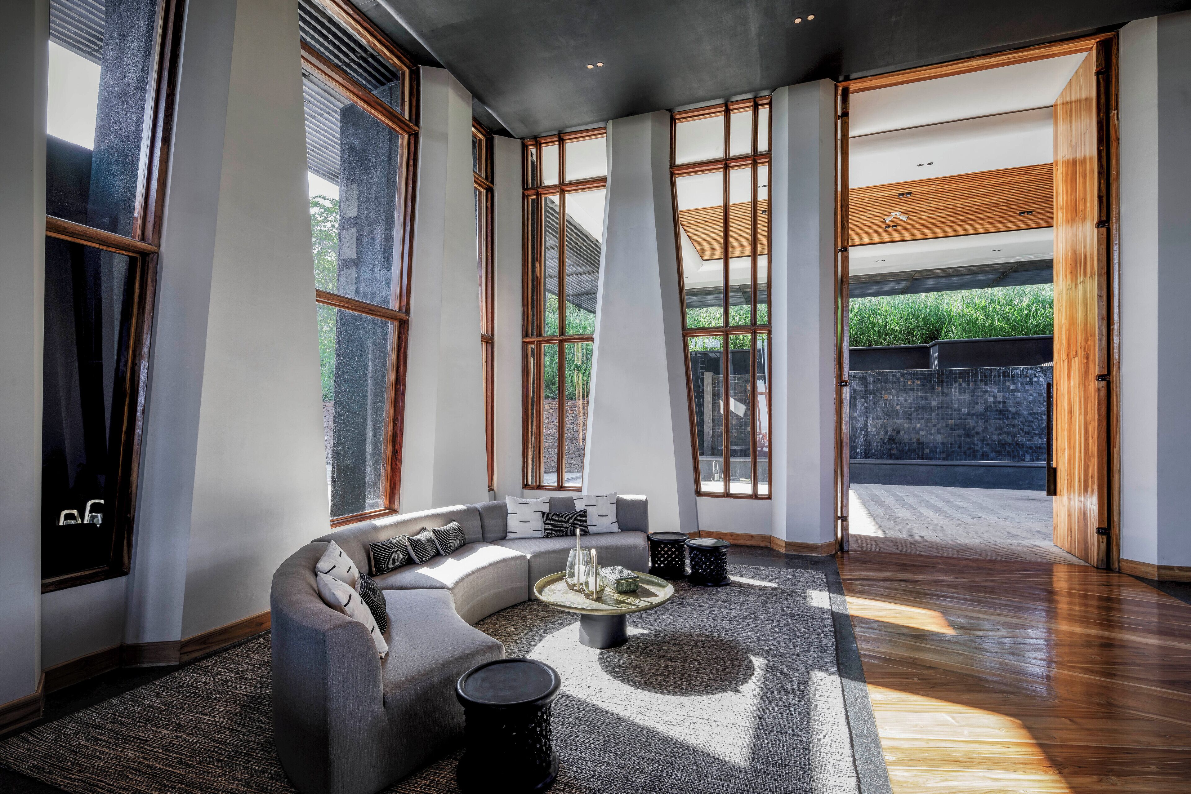 A modern living room with a curved sectional sofa, a coffee table, and large windows offering views of greenery.