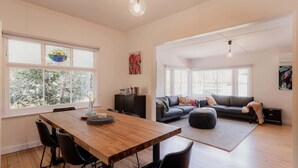 A spacious living area with a wooden dining table, black chairs, a gray sofa, and a black ottoman. Large windows provide natural light and views of greenery outside. - Treelined retreat Lenah Valley (Lenah Valley)