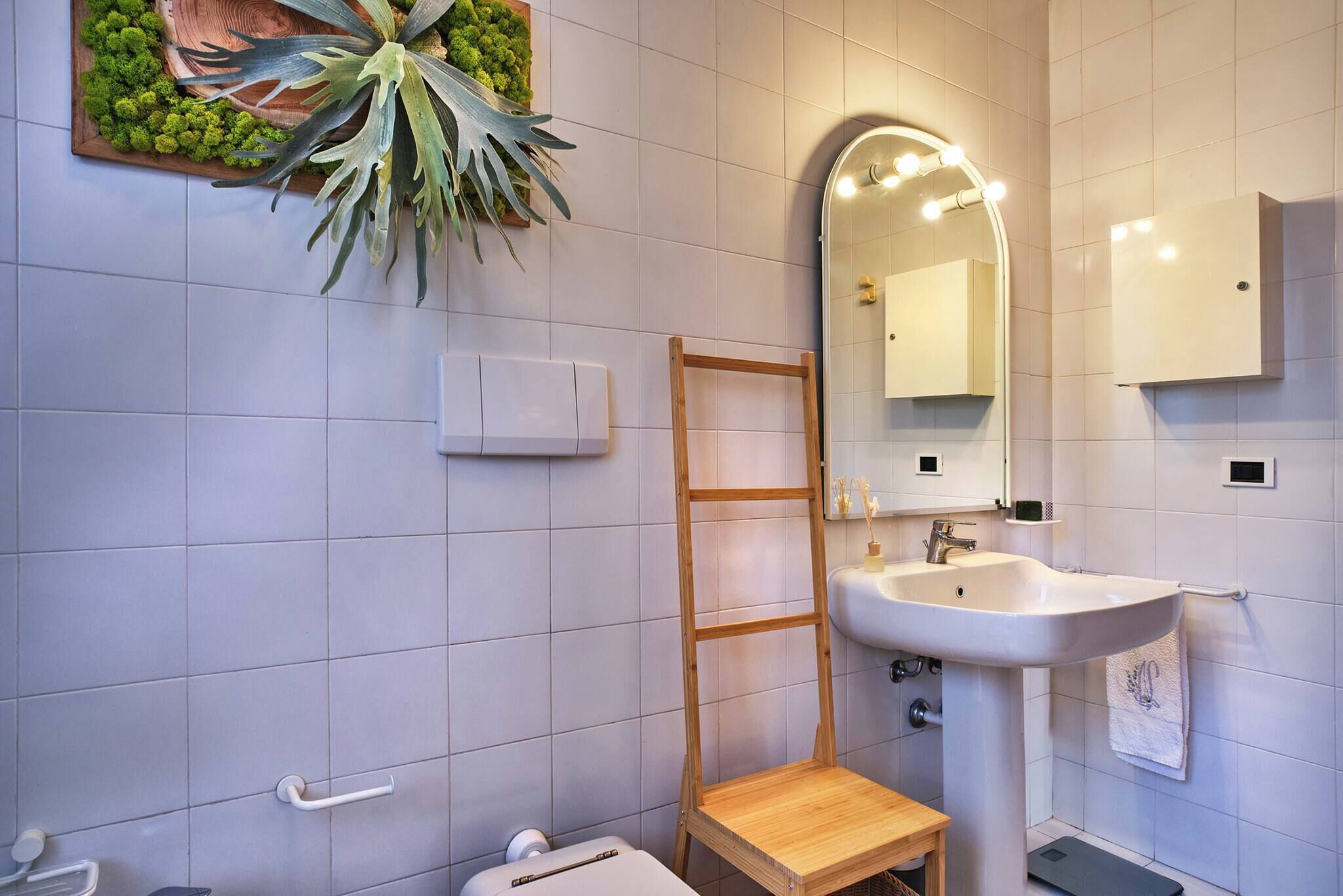 A bathroom with a wooden ladder, a sink, a mirror with lights, and a wall-mounted plant.
