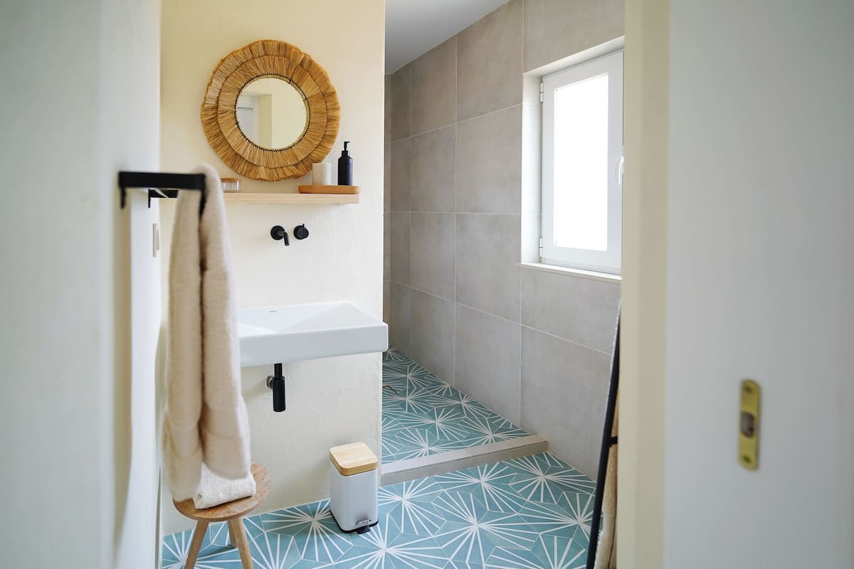Une salle de bain avec un lavabo blanc, un miroir rond, un porte-serviettes, une petite poubelle et une fenêtre.