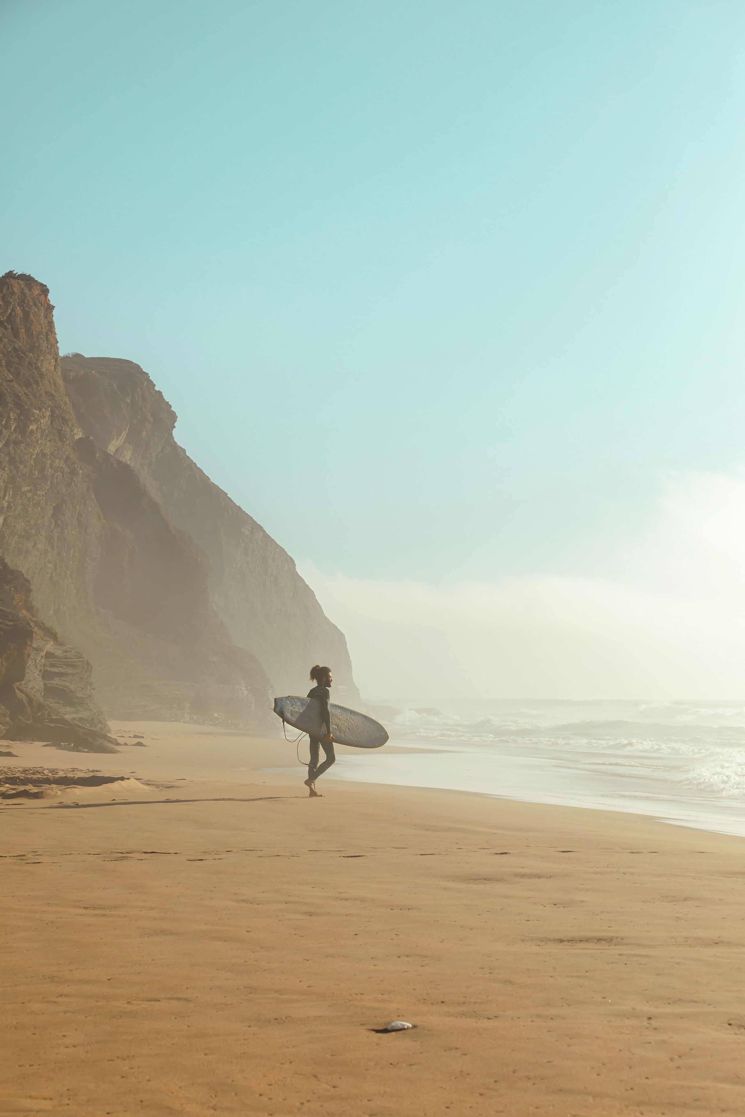 Eine Person geht über einen sandigen Strand, ein Surfbrett tragend, mit Klippen im Hintergrund und blauem Himmel.