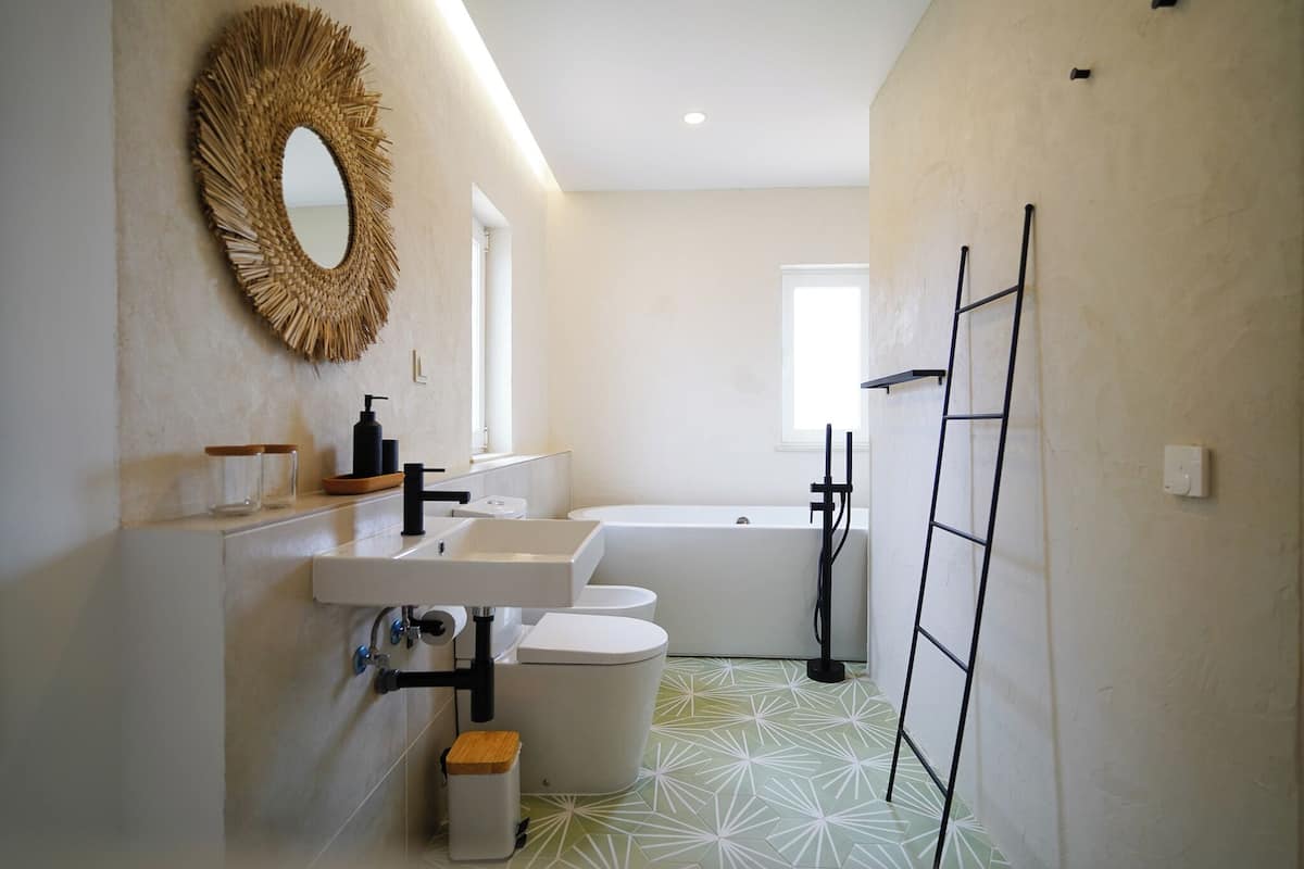Une salle de bain avec un lavabo, des toilettes et une baignoire blancs, un miroir à cadre en bois et un carrelage décoré.
