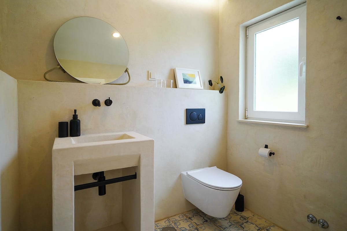 Une salle de bain avec un lavabo blanc, des toilettes, un miroir, un tableau encadré et une fenêtre.