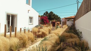 A narrow pathway flanked by tall grasses and flowering plants, leading to a white building with multiple windows and a door.