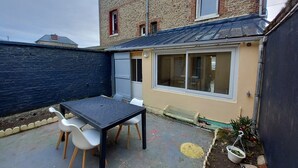 Un patio con una mesa y sillas, un pequeño jardín con plantas, y un edificio de fachada de ladrillo y ventanas.