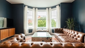 A modern living room with a leather sofa, a glass coffee table, and a TV on a wooden cabinet. Large windows with white curtains offer a view of greenery and buildings outside.