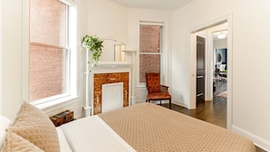 A bedroom with a large bed, a fireplace, a chair, and a view of a dining area.