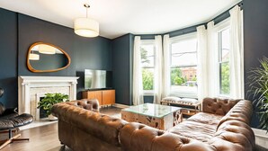 A living room with a dark blue wall, a white fireplace, a leather sofa, a glass coffee table, and a television on a wooden cabinet.