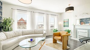 Een moderne woonkamer met een witte bank, een glazen salontafel en een groene fauteuil. Er is een open haard met een witte schouw en een schilderij erboven.