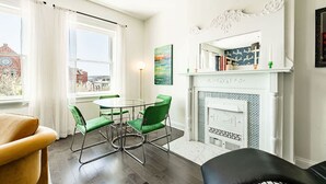 Een moderne woonkamer met een open haard, groene stoelen, een ronde tafel en een stadszicht door grote ramen.