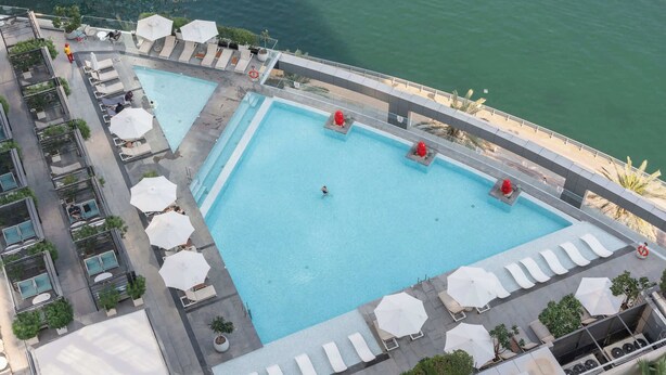 Une grande piscine rectangulaire dotée de plusieurs espaces de détente et de parasols, entourée d’un immeuble de grande hauteur offrant une vue sur l’eau.
