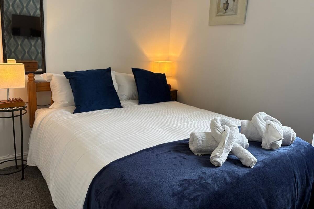 A neatly made bed with a white duvet and blue pillows, a bedside lamp, and a framed picture on the wall.