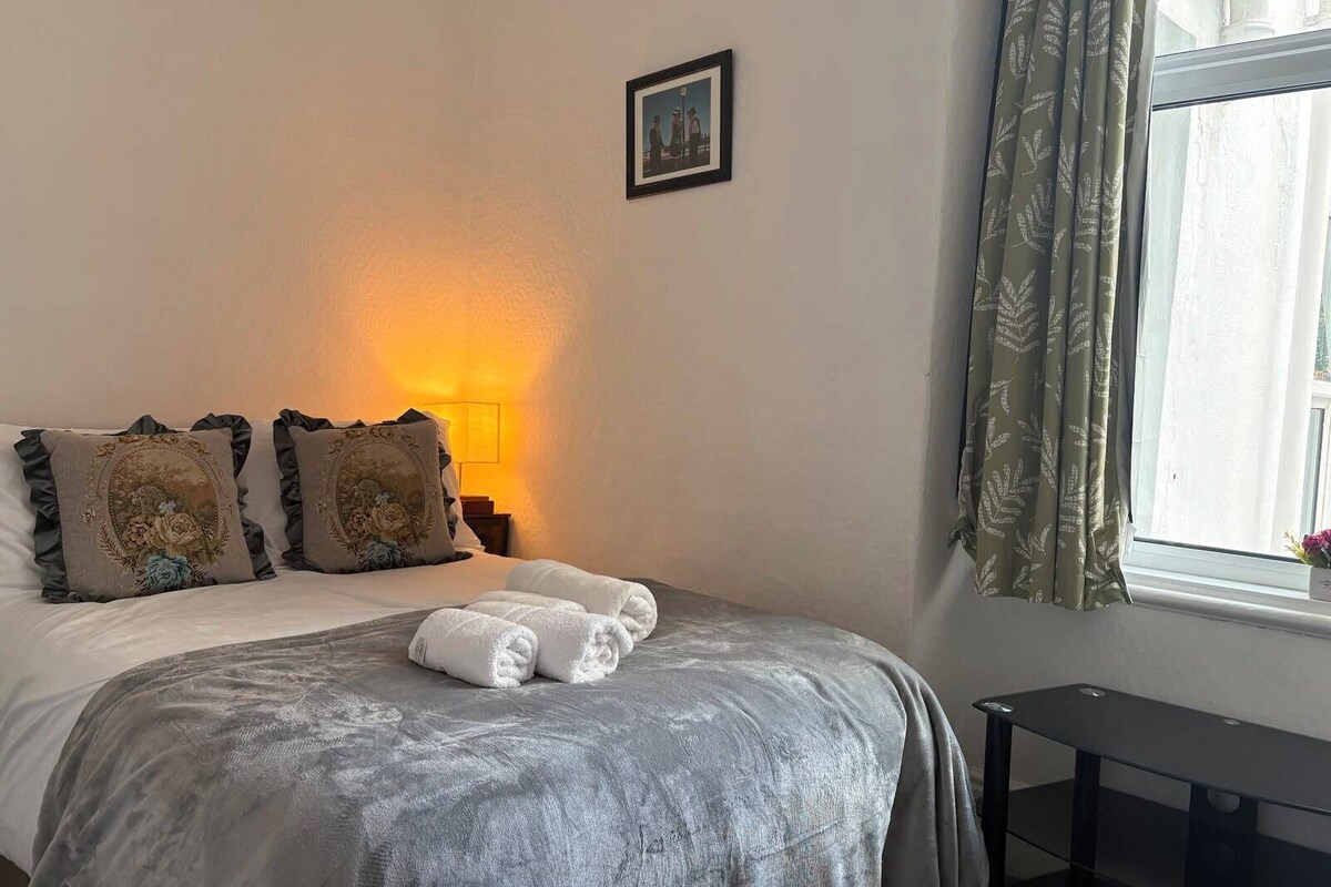 A hotel room with a bed, pillows, a bedside lamp, a window with curtains, and a small table.