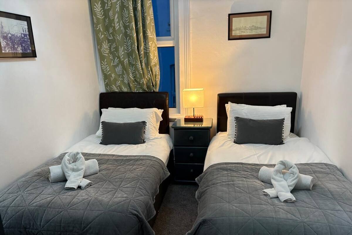 A hotel room with two beds, each with a dark quilted bedspread and white pillows. There are towels folded on the beds, and a nightstand with a lamp between the beds.