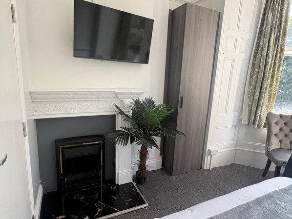 A room with a fireplace, a television mounted above it, a potted plant, and a chair by the window. - Athol House (Douglas)