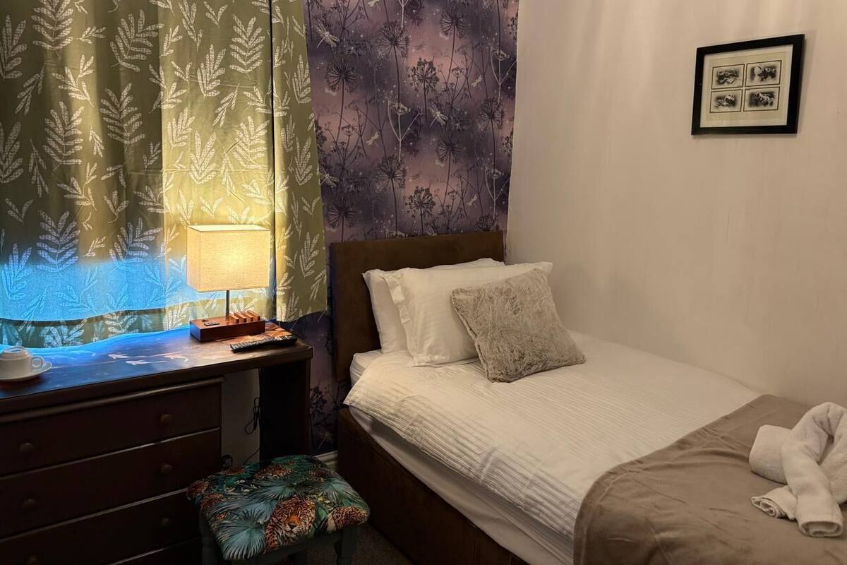 A neatly arranged bedroom with a bed, a bedside table with a lamp, a small stool, and a framed picture on the wall.