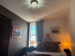 A hotel room with a bed, a nightstand, a lamp, a window with curtains, and two framed pictures on the wall. - Athol House (Douglas)