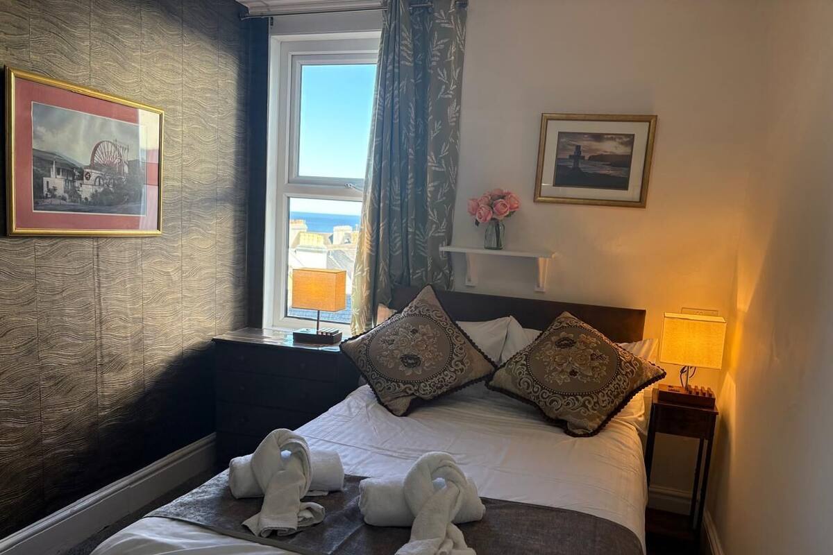 A hotel room with a bed, pillows, a nightstand, a lamp, and a window with curtains.