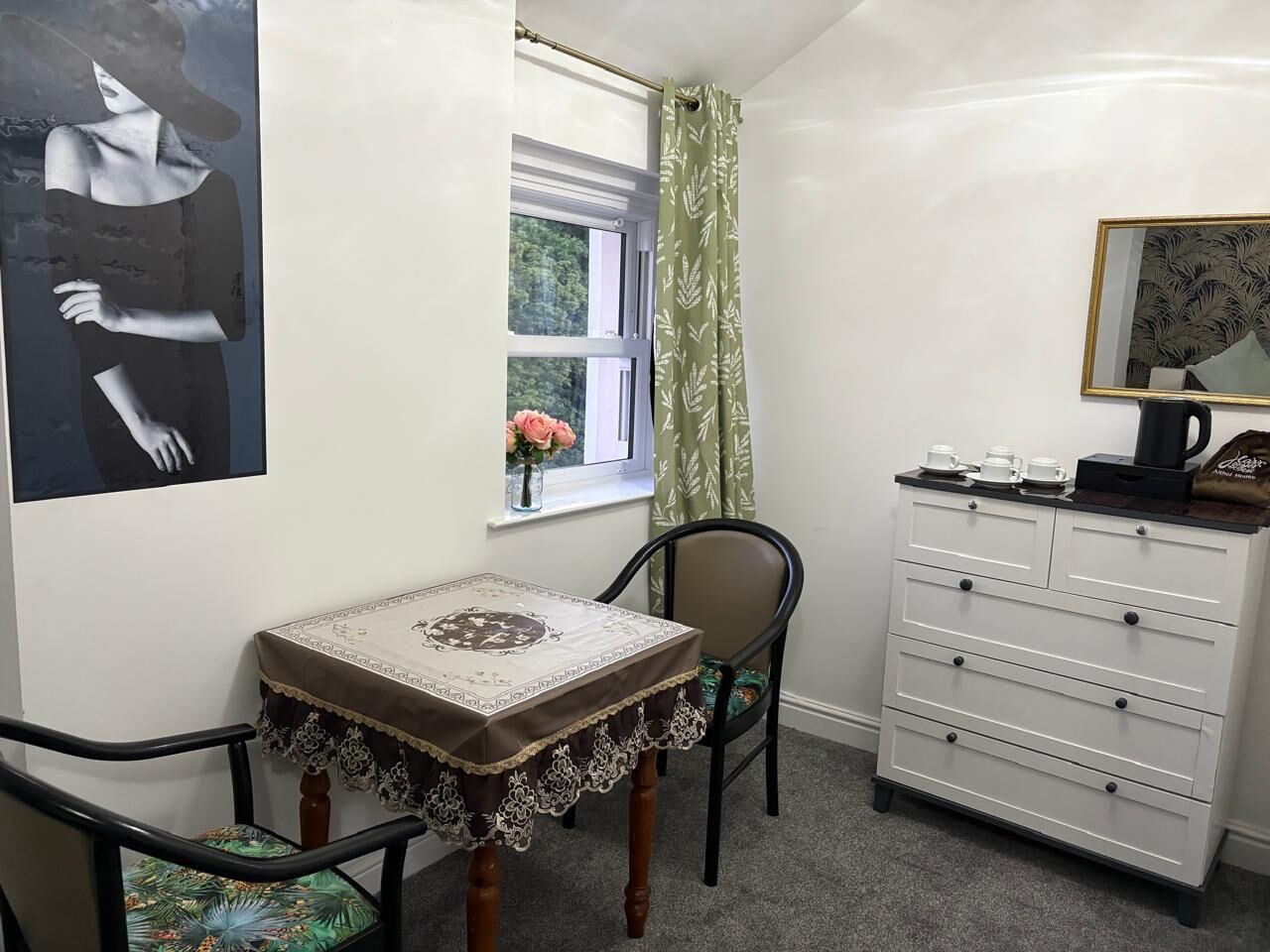 Una habitación pequeña con una mesa, una silla, un aparador, una ventana con cortina y un cuadro en la pared.