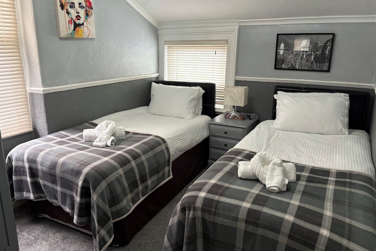 A hotel room with two beds, each with a plaid blanket and white pillows. There is a nightstand with a lamp and a small table with a bag on it. The room has a window with blinds and a painting on the wall.