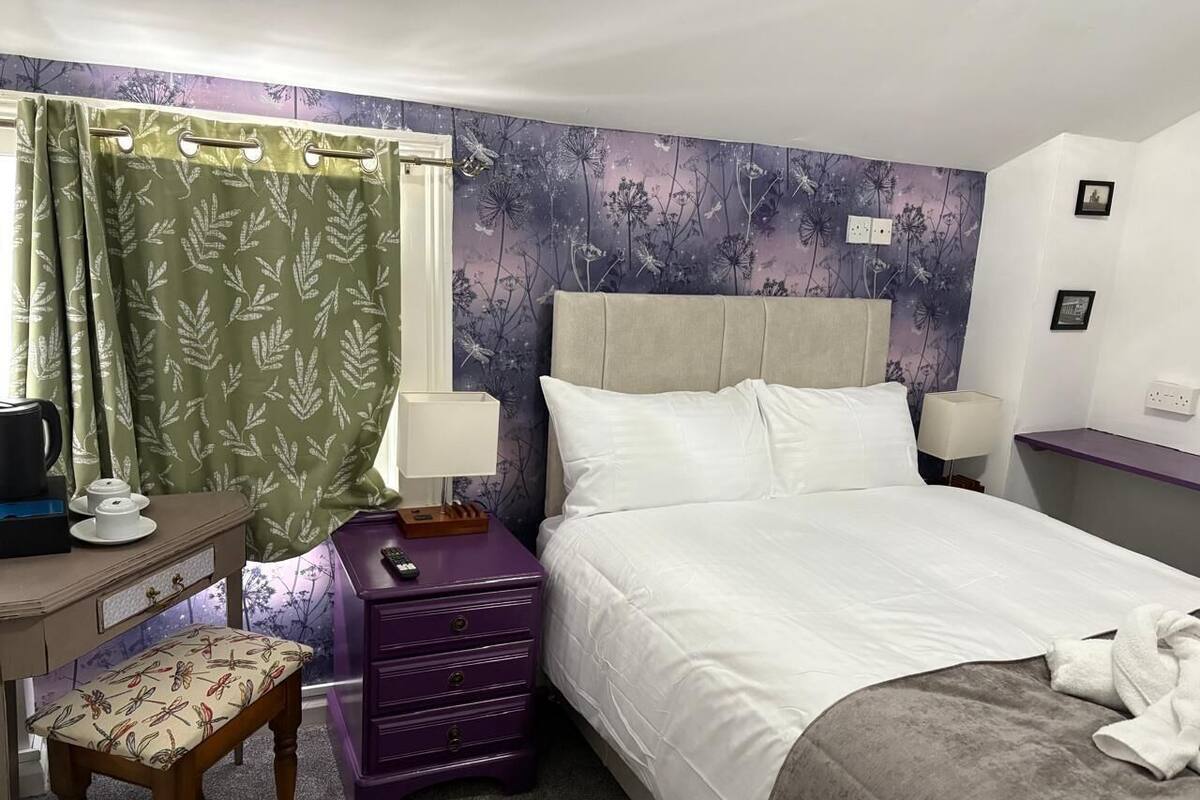 A hotel room with a bed, a desk with a chair, a purple dresser, a green curtain, and a floral wallpaper.