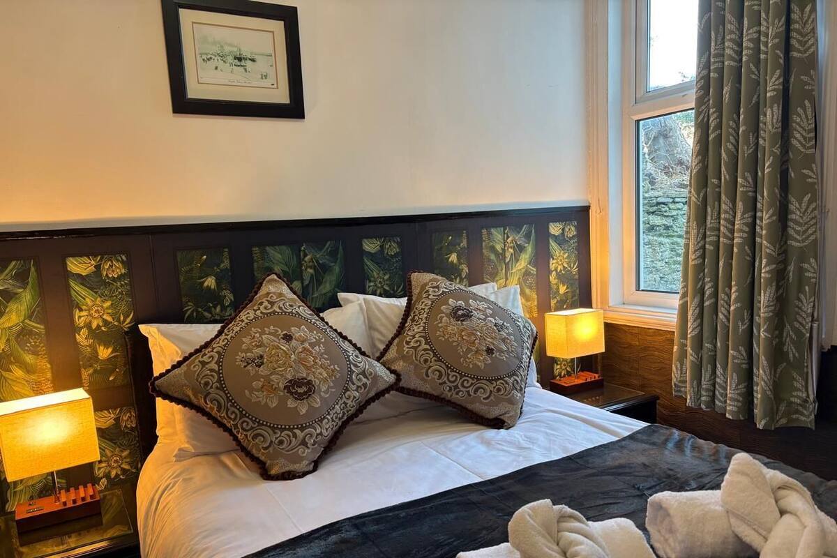A hotel room with a large bed, two decorative pillows, a nightstand with a lamp, and a window with curtains.
