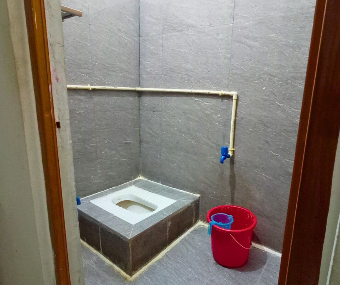 Een kleine betegelde badkamer met een toilet, een rode emmer en een blauwe kraan.