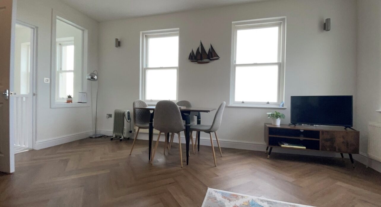 Een ruime woonkamer met een houten vloer, een eethoek met tafel en stoelen, een televisie op een houten kast, en grote ramen die zorgen voor natuurlijk licht.