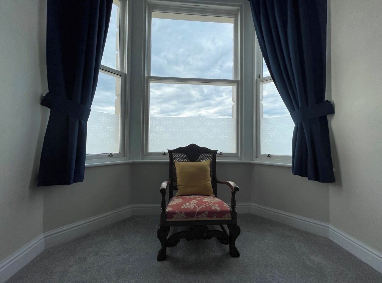 A corner room with a large window, a dark wooden chair with a cushion, and a carpeted floor.