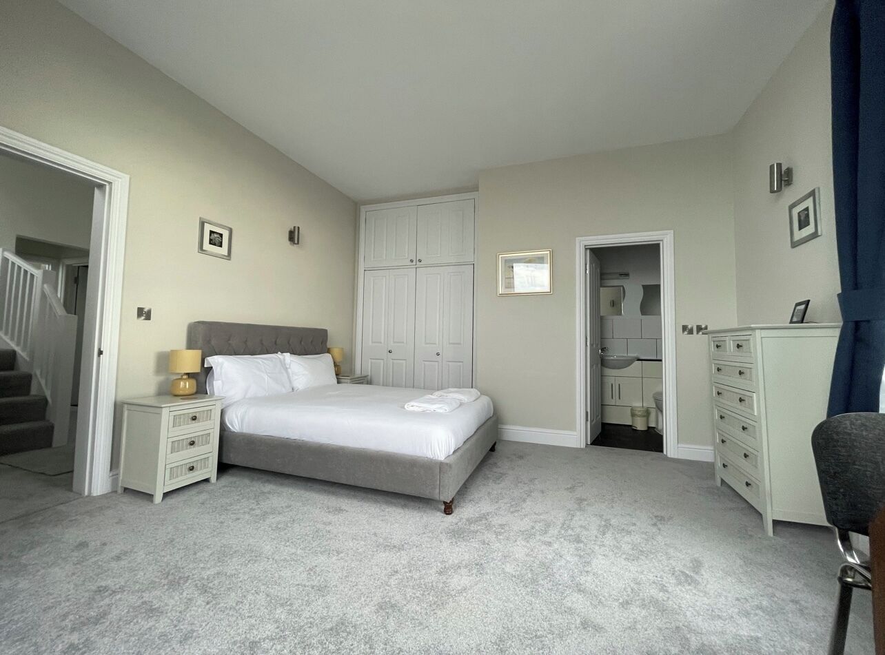 Quarto com cama, mesas de cabeceira, cômoda e banheiro visível por uma porta aberta.