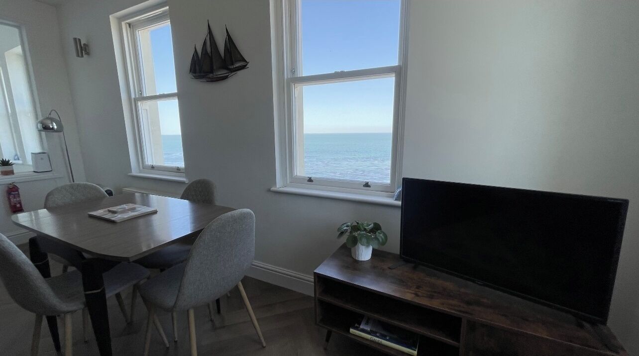 Ein Zimmer mit Esstisch, Stühlen, Fernseher und Meerblick durch große Fenster.