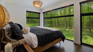 A modern bedroom with a large bed, a hanging light fixture, and a view of a forest through large windows. - Eastman - Spa - Zénitude (Eastman)