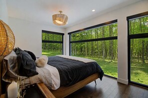 A modern bedroom with a large bed, a hanging light fixture, and a view of a forest through large windows. - Eastman - Spa - Zénitude (Eastman)
