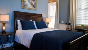 A hotel room with a bed, two bedside lamps, a painting on the wall, and a window with curtains. - Kinder Rm, The Lodge at  Old City Hall (Jefferson City)