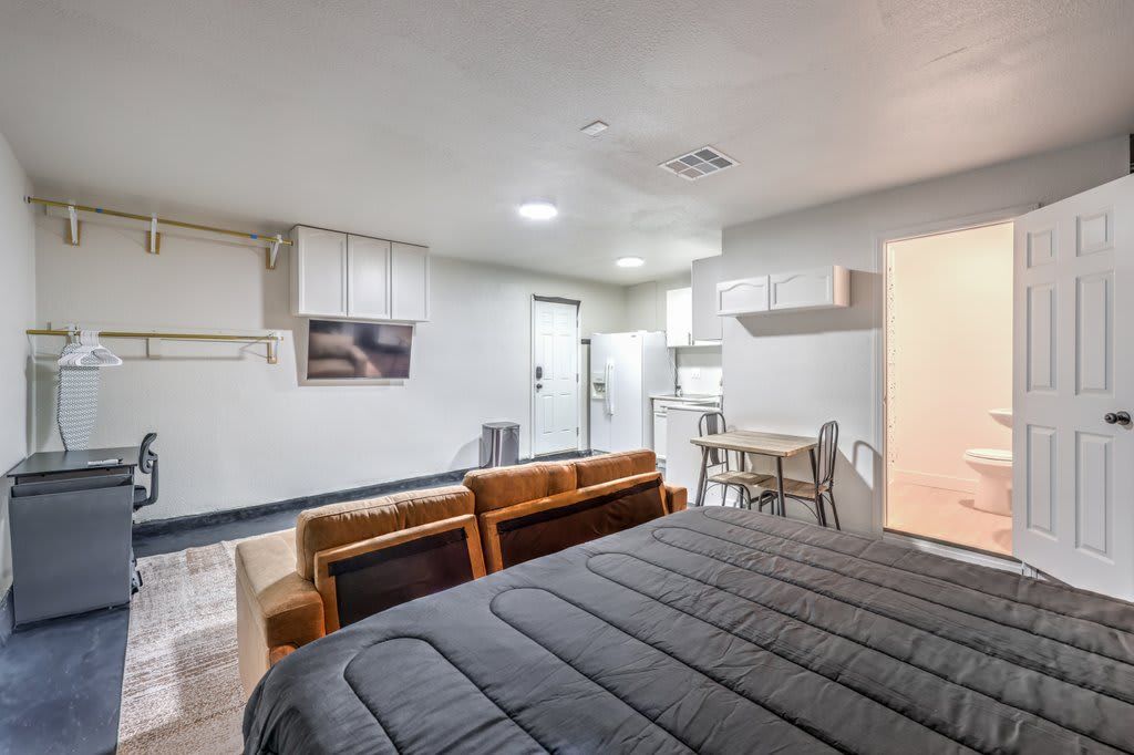 A bedroom with a bed, a desk, and a chair. There is a kitchenette with a refrigerator and a sink.
