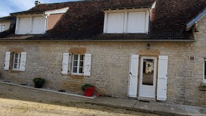 Un bâtiment en pierre doté de trois fenêtres, chacune ornée de volets blancs, et d’une porte surmontée d’un petit auvent.