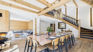 Een ruime eethoek met een lange houten tafel gedekt voor zes personen, omringd door stoelen. Een trap met houten leuningen leidt naar een hoger gelegen verdieping.