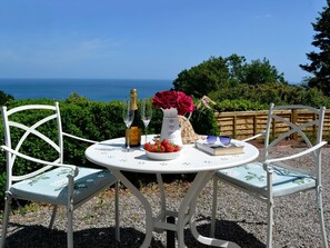 Une table ronde d’extérieur entourée de chaises blanches, une bouteille de champagne, un vase rempli de fleurs rouges, un livre et un saladier de fraises.