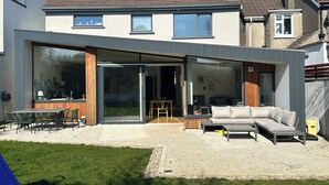 A modern house with a large glass door leading to a patio area, a dining set, and a sofa. - Bright Spacious 5-bedroom house in Dundrum with sunken Trampoline and Treehouse. (Dublin 8)