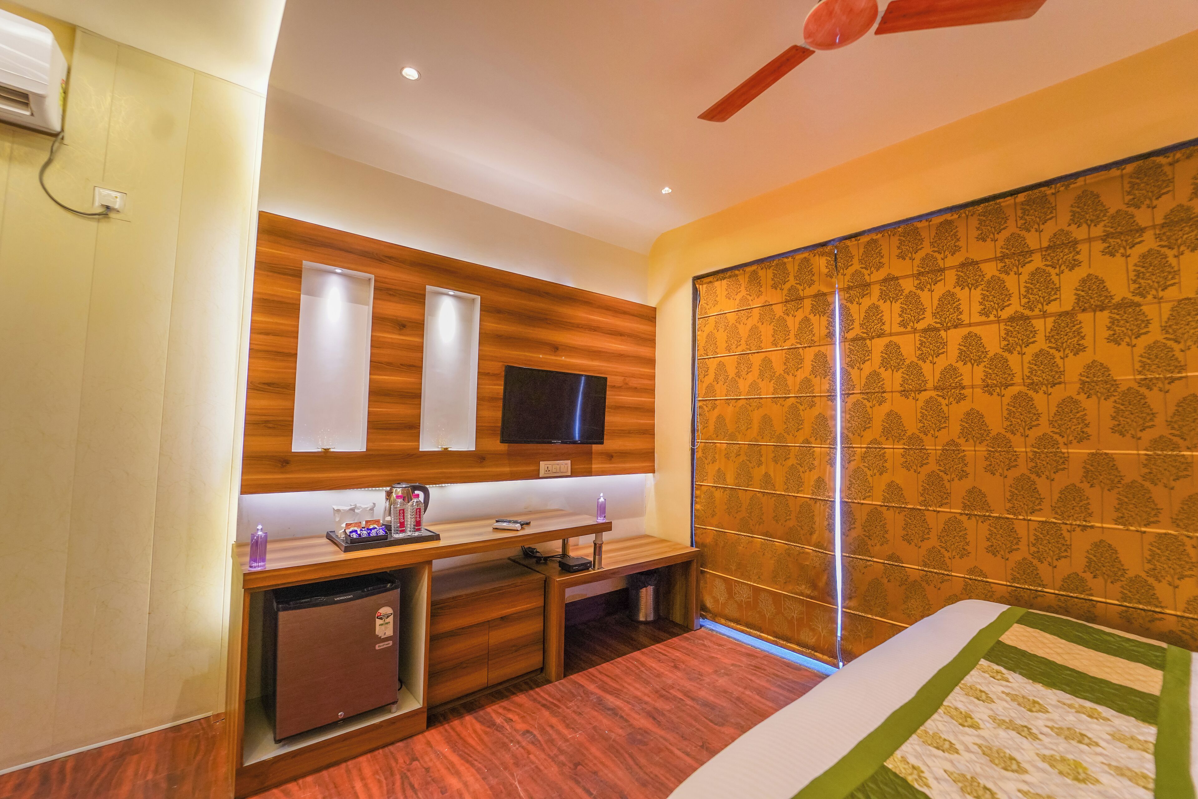 A hotel room with a wooden desk, a television, a small refrigerator, and a bed with a quilt.