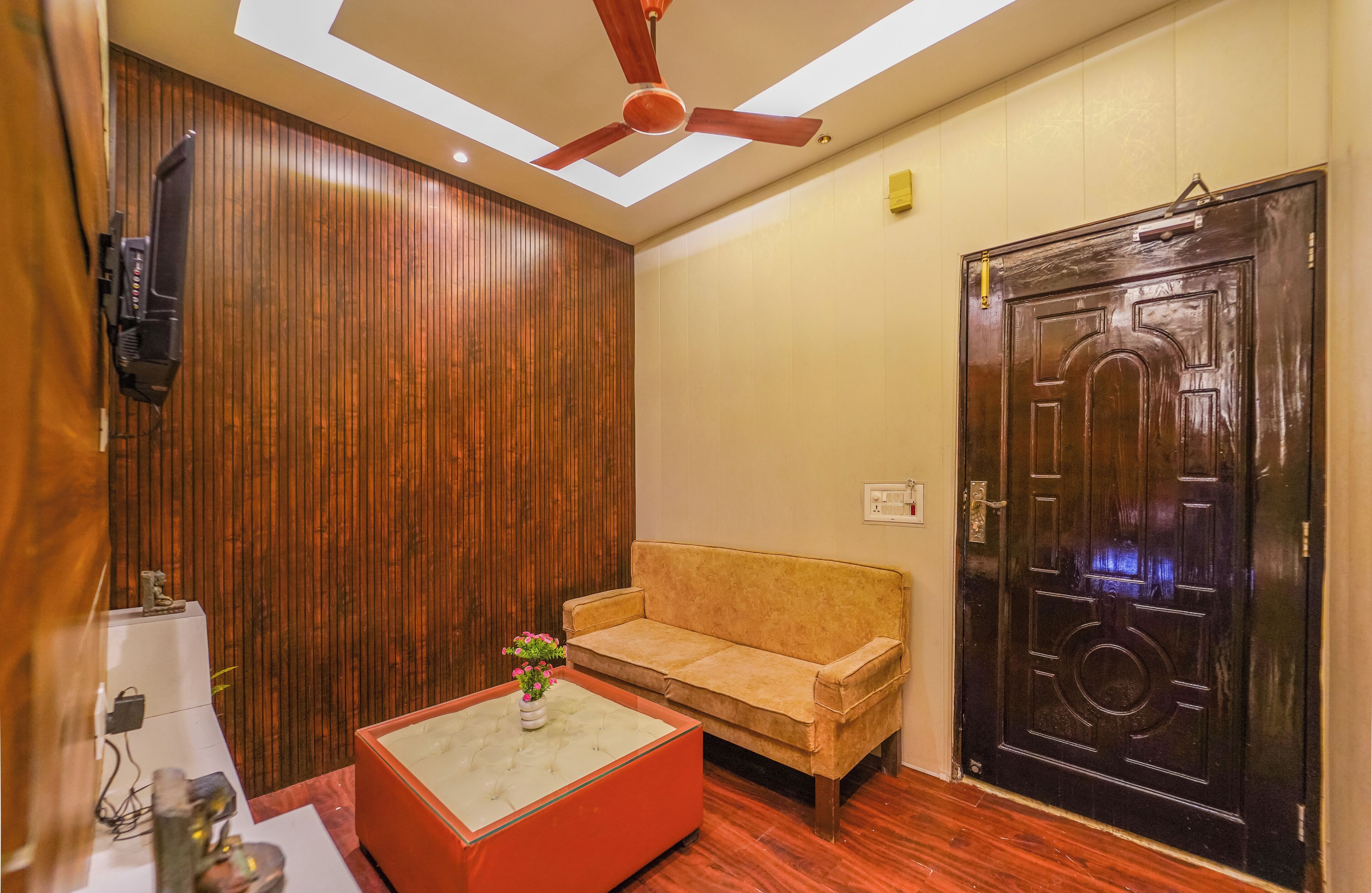 Quarto com parede de painel de madeira, sofá bege, mesa de centro vermelha e porta preta com detalhe decorativo.