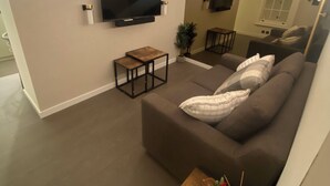 A modern living room with a large flat-screen TV mounted on the wall, a dark brown sofa, a small wooden coffee table, and a potted plant.